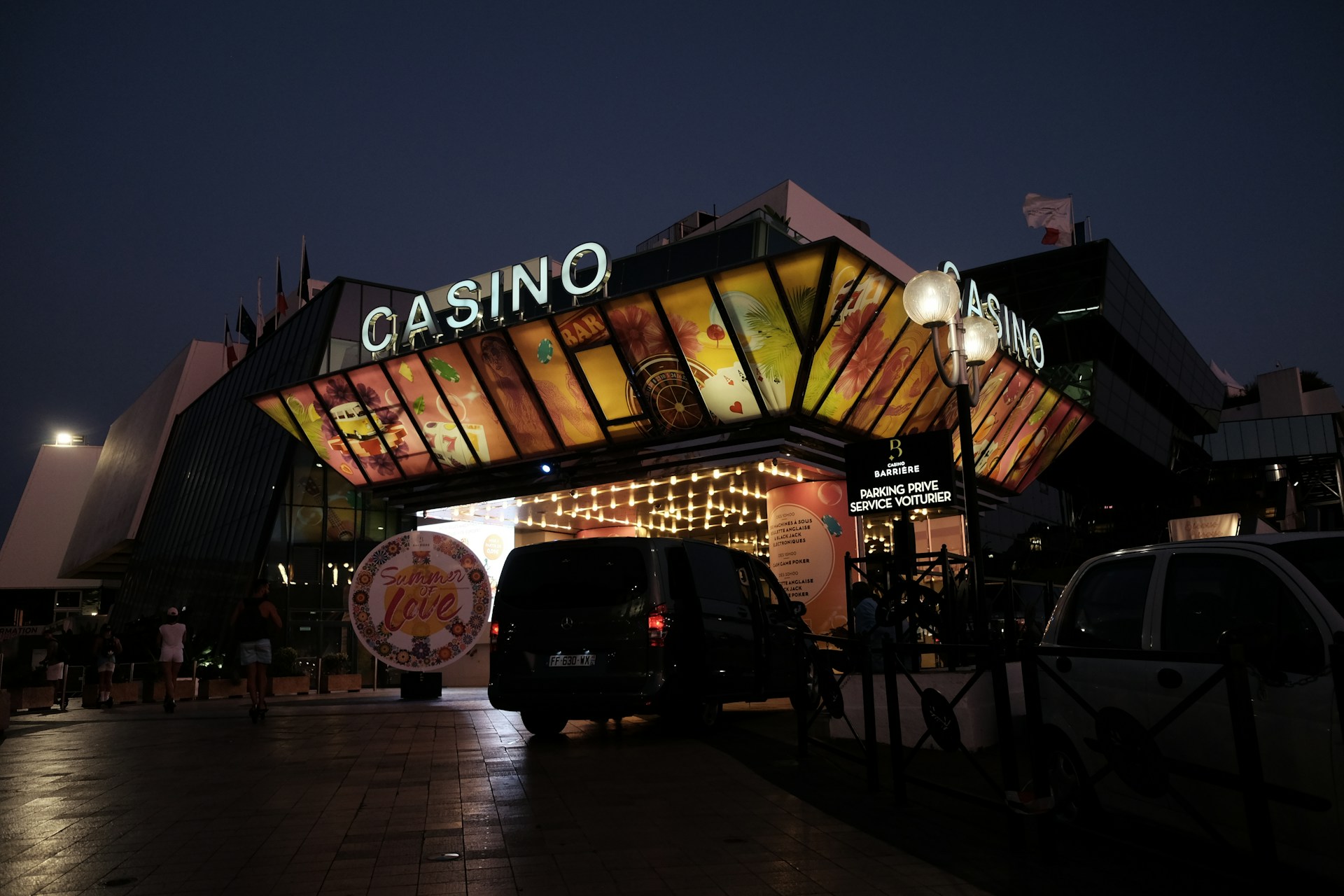 A building with a brightly illuminated entrance is seen at night. The sign above reads 'Casino' in bright letters. The facade features colorful graphics depicting various indulgent themes. Several vehicles are parked near the entrance, and people are visible in the vicinity. Tall lamp posts with spherical lights are present, casting a warm glow.